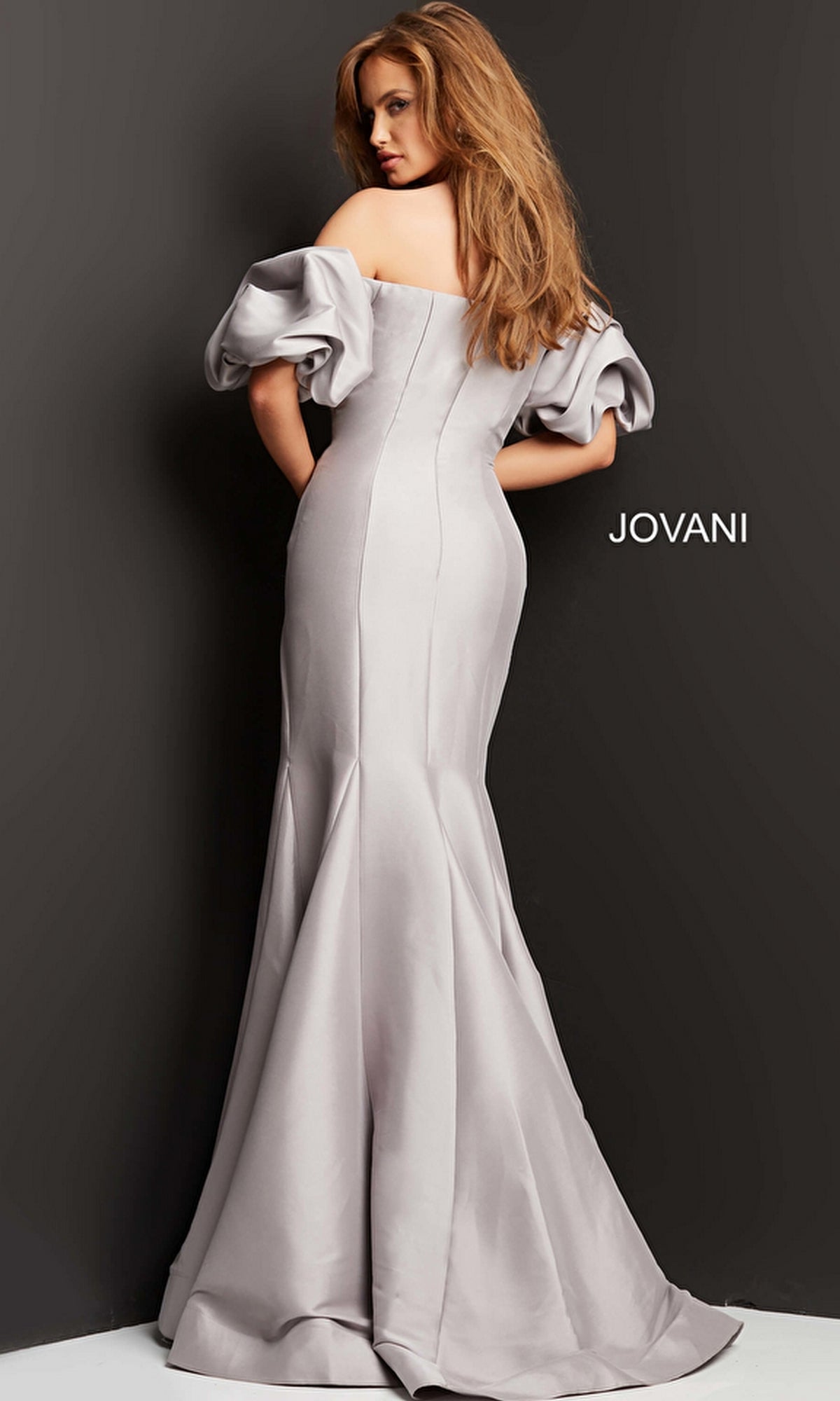 A woman poses with her back to the camera against a dark background, wearing the Jovani 08361 Puff-Sleeve Mikado Evening Dress in silver, featuring off-the-shoulder puffed sleeves and a mermaid silhouette. The word